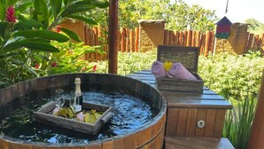 Un bain à remous en bois, avec deux verres et une bouteille, entouré d’une végétation luxuriante et d’une terrasse en bois.