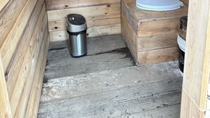 A small wooden cabin with a single white toilet, a metal trash can, and a shelf against the wall. - Enjoyable 1-bedroom RV in Seward (Seward)
