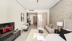Quarto de hotel moderno com televisão de tela plana, sofá, cama, pequena mesa de jantar e área de cozinha ao fundo.