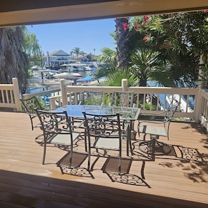 Ein Holzbalkon mit Tischplatte aus Glas und Metallstühlen, mit Blick auf eine Marina mit Booten und Palmen.