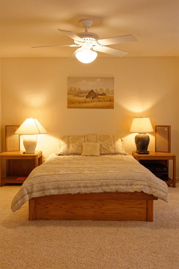 A bedroom with a wooden bed, two bedside tables with lamps, a ceiling fan, and a painting on the wall.