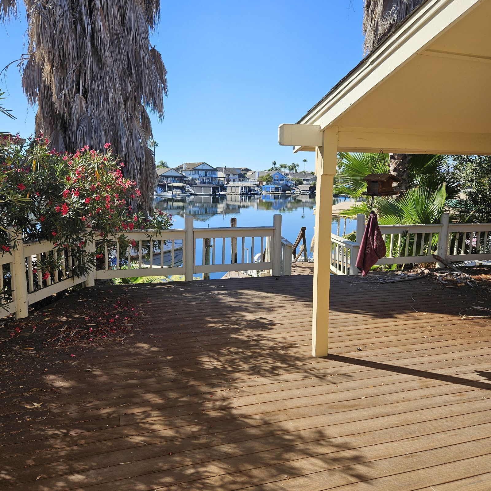Une terrasse en bois avec une balustrade blanche, donnant sur une marina où l’on aperçoit des bateaux et des maisons au loin.