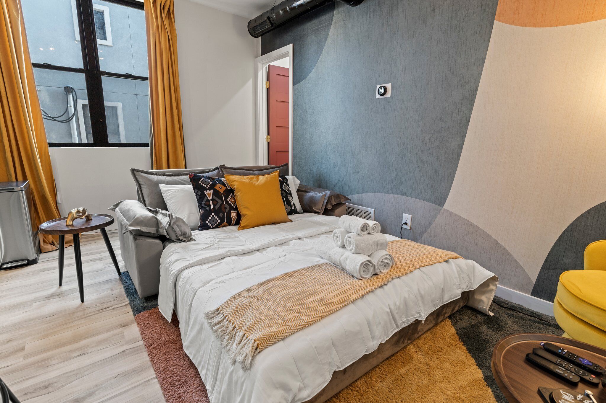 Habitación de hotel moderna con una cama grande, una mesita auxiliar pequeña, un sillón amarillo y vistas a la calle a través de la ventana.