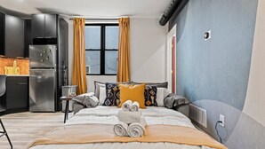 Ett modernt hotellrum med en stor säng, ett litet köksutrymme och ett fönster med gardiner.