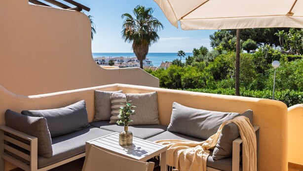 A sunlit outdoor seating area with a sofa, cushions, a table, and a large umbrella.