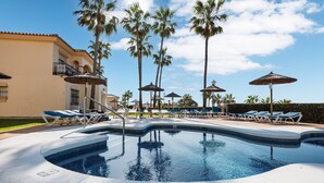 A swimming pool with lounge chairs and umbrellas, surrounded by palm trees and a building with balconies.