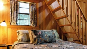 A wooden room with a bed, a window with curtains, a lamp, and a wooden staircase.