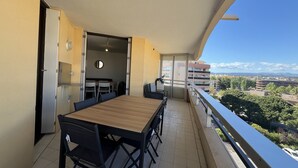 Un balcón con mesa y sillas de madera, una puerta corredera que da a un comedor, y vistas a edificios y zonas verdes.