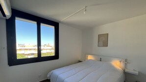 Un dormitorio con cama, mesita de noche, luz adosada a la pared, ventana con vista a edificios y un cuadro enmarcado.