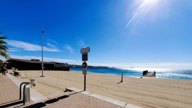 Una playa con un paseo marítimo pavimentado, un cartel público y un cielo azul despejado.