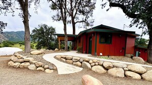 Uma cabana vermelha com um caminho de pedra que leva à entrada, rodeada por pedras grandes e árvores.