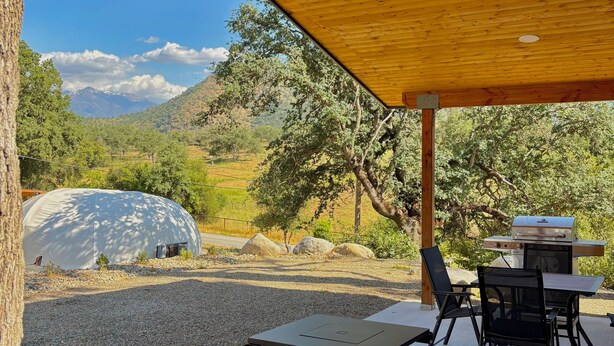 Área externa coberta com mesa, cadeiras, churrasqueira e vista para montanha e árvores.