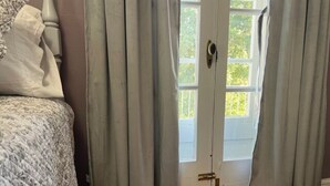 A room with a bed, a door with curtains, and a view of greenery outside. - The Azalea Room w/ Queen Bed (Woodsfield)