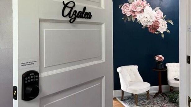 A room with a white door labeled "Azalea," a dark blue wall with a floral painting, two white chairs, and a small table with a vase. - The Azalea Room w/ Queen Bed (Woodsfield)