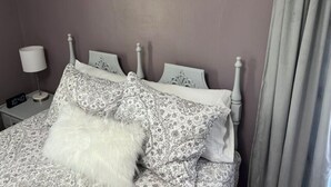 A bed with patterned bedding and a fluffy pillow, a bedside table with a lamp, and a clock. - The Azalea Room w/ Queen Bed (Woodsfield)