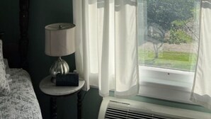Une chambre avec un lit, une table de chevet avec une lampe, une fenêtre avec des rideaux blancs et une vue sur la verdure à l’extérieur.