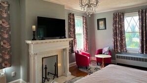 Une chambre à coucher avec une cheminée, une télévision, un lit, un fauteuil rouge et une fenêtre avec des rideaux.