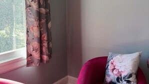 Une pièce avec un rideau à motifs floraux, un tableau encadré au mur et un fauteuil rouge agrémenté d’un coussin à motifs floraux.