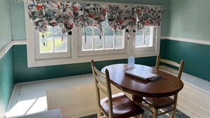 Un comedor con una mesa redonda de madera y cuatro sillas, una ventana con cortina floral y paredes azul verdoso.