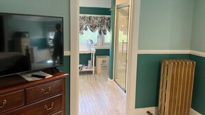 Una habitación con paredes verde azulado, un aparador de madera con cajones, un televisor, un radiador y una puerta que da a otra habitación.