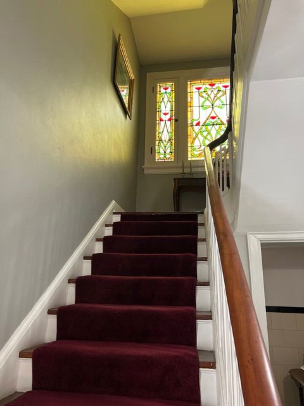 Una escalera con peldaños alfombrados de rojo y barandilla de madera, que conduce a una habitación con un ventanal de vitral.