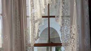 A church interior with a wooden cross, white lace curtains, and a stone bench.