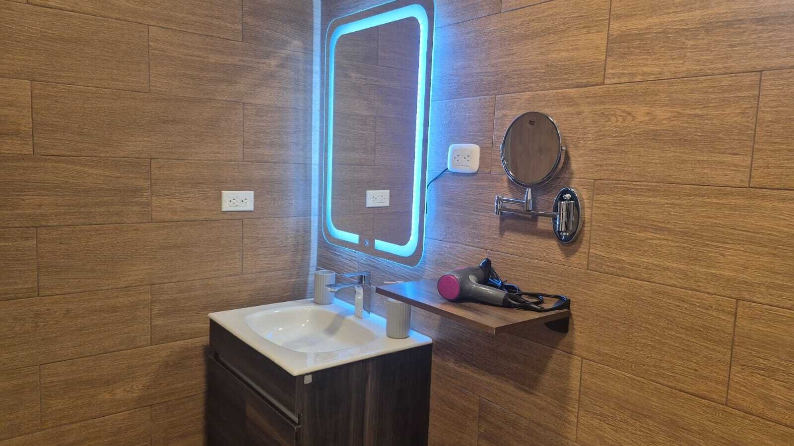 Un bany modern amb terra de fusta, un lavabo blanc, un mirall amb llum de neó i un mirall rodó, i un assecador de cabells sobre una prestatgeria.