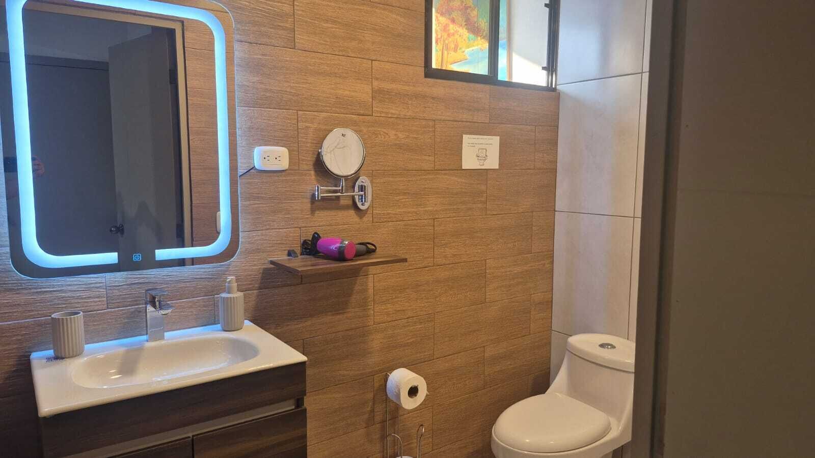 Ein modernes Badezimmer mit einem Holzwaschtisch, einem Spiegel mit integrierter Beleuchtung, einer Toilette und einem Ablagefach für Pflegeutensilien.