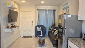 A compact living space with a door, a seating area with a sofa and a chair, a small table, a refrigerator, and a television mounted on the wall.