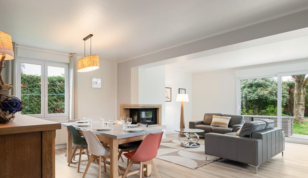 En moderne stue med sofa, spisebord og stole. Der er en pejs og et stort vindue med udsigt til grønne omgivelser.
