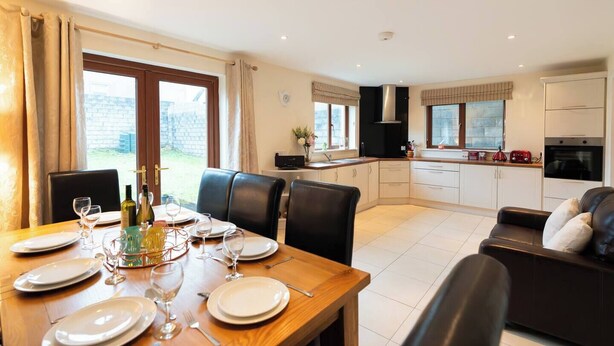 A modern kitchen with white cabinets, a dining area with a wooden table set for four, and a black leather sofa.