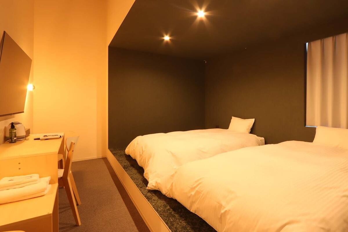Ein Hotelzimmer mit zwei Betten, einem Schreibtisch mit Stuhl, einem Flachbildfernseher und einem Fenster mit Vorhängen.