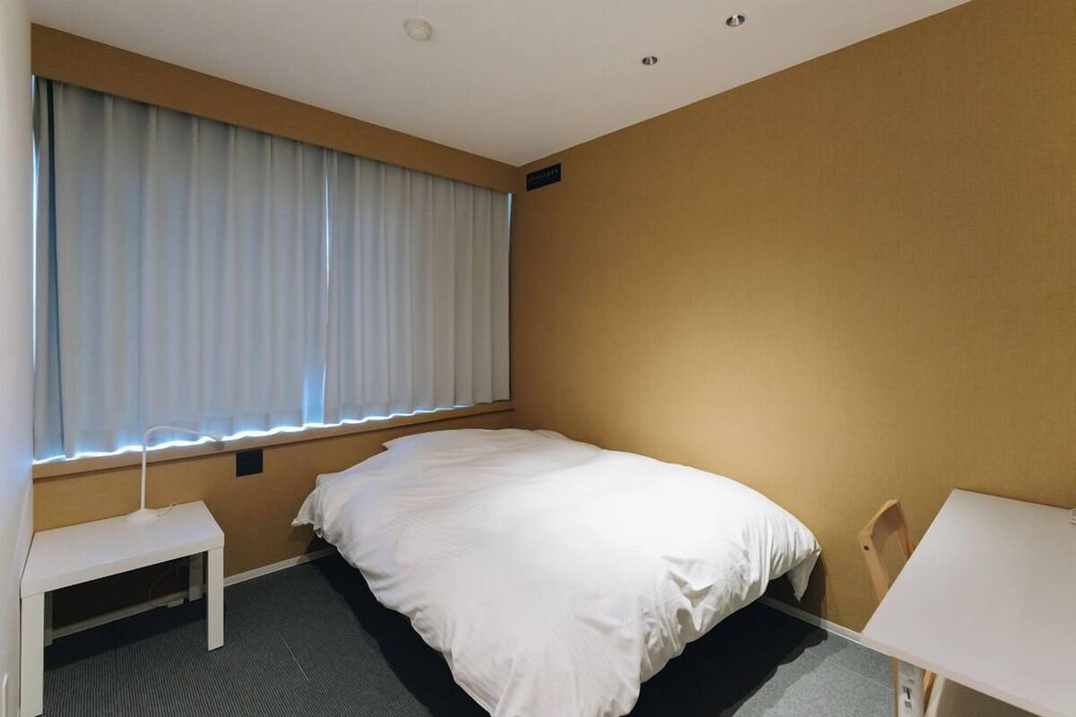 Ein Hotelzimmer mit einem Einzelbett, einem kleinen Schreibtisch, einem Stuhl und einem fenster mit Vorhängen.