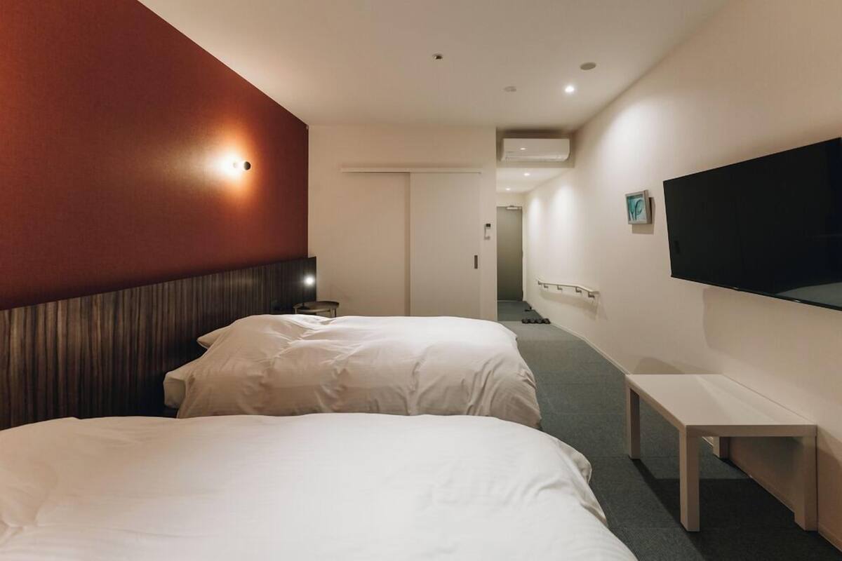 Ein Hotelzimmer mit zwei Betten, einem Flachbildfernseher an der Wand, einem Nachttisch und einer Bank.