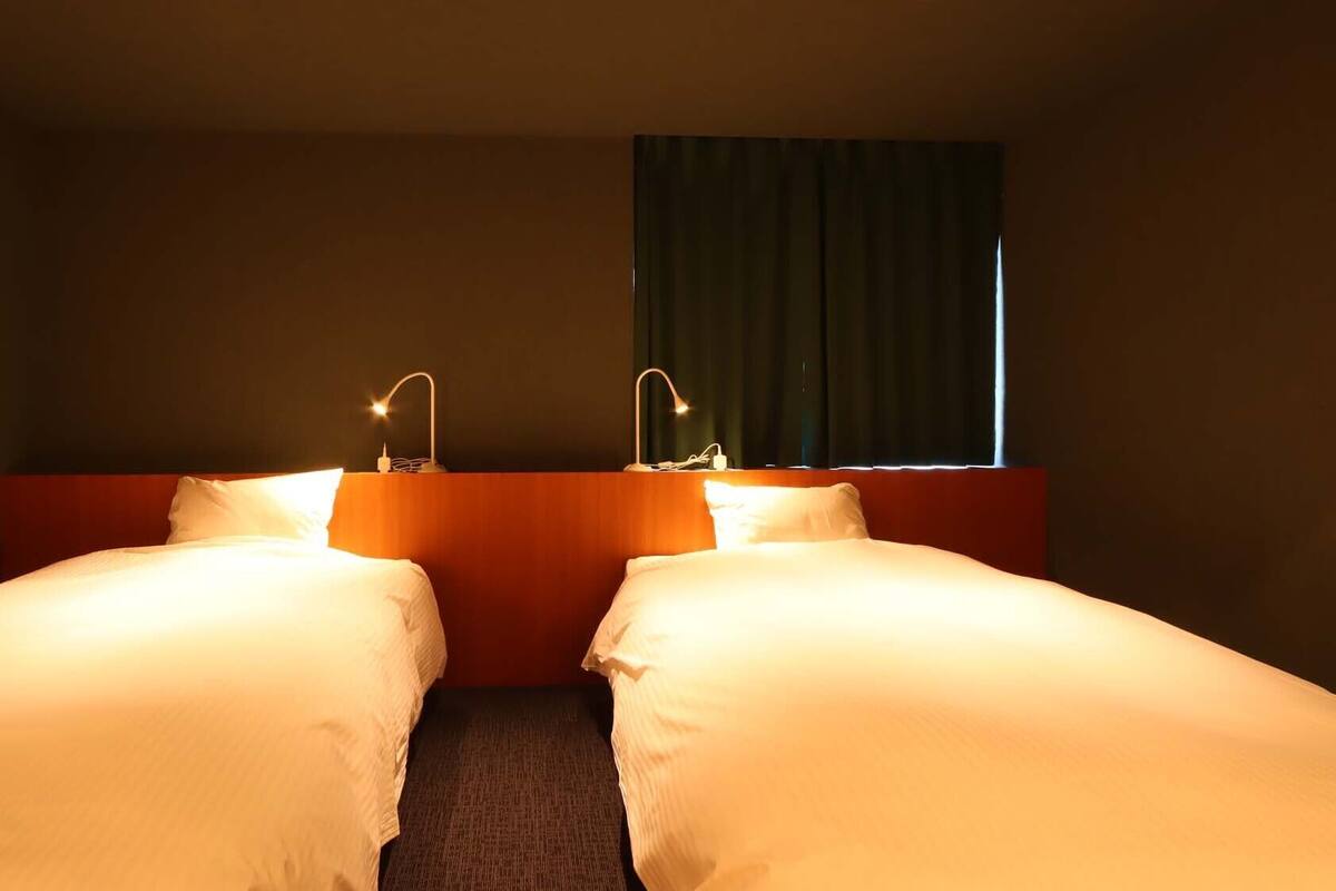 Ein Hotelzimmer mit zwei Einzelbetten, einem Holzkopfende, einem Fenster mit Vorhängen und Nachttischlampen.