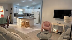 Een moderne woonkamer met een bank, een glazen salontafel en een roze fauteuil. Er is een keuken met witte kasten en een grote televisie aan de muur bevestigd.