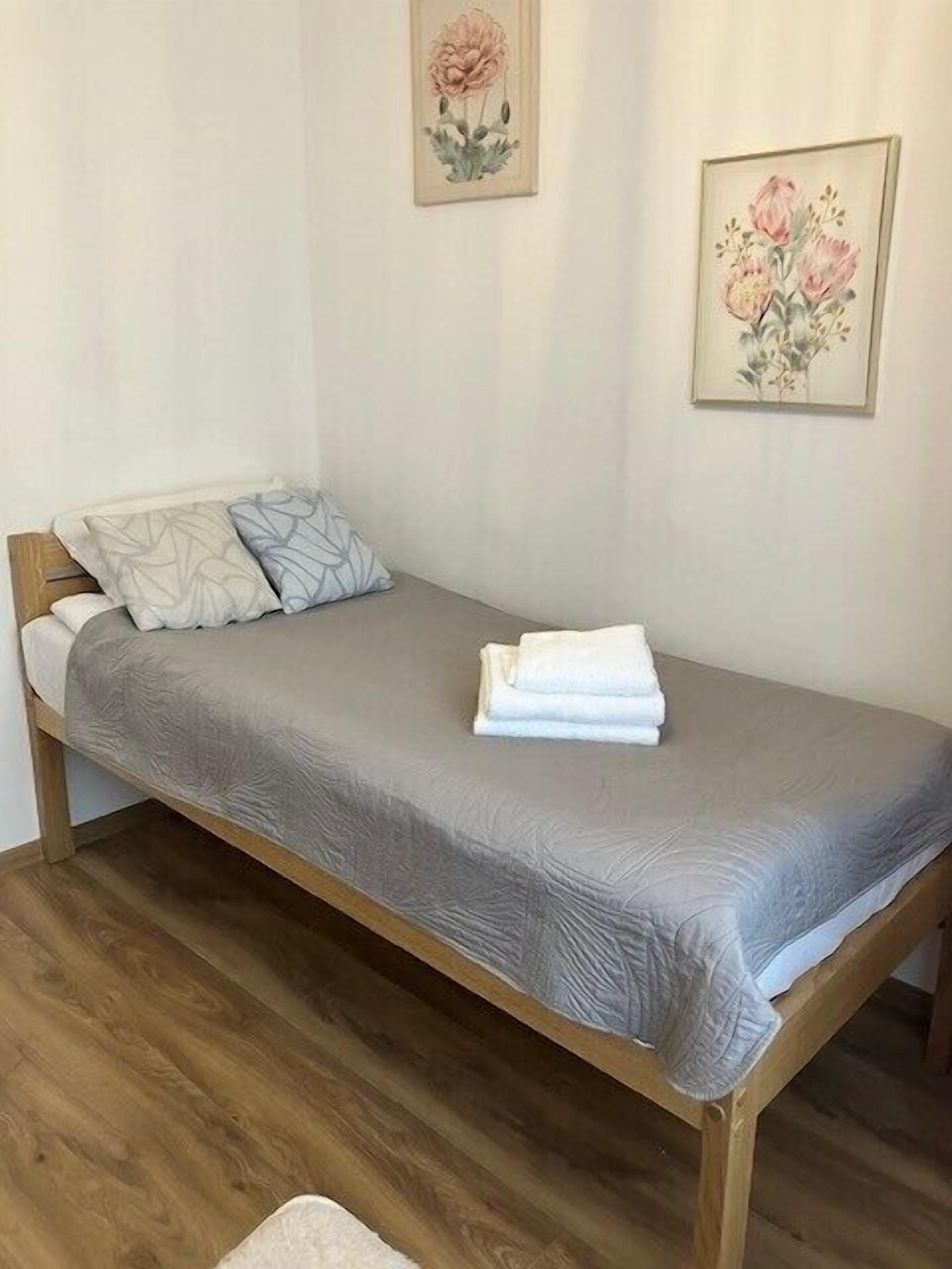 A single bed with a grey bedspread and two pillows, a folded towel on the bed, wooden flooring, and two framed floral paintings on the wall.