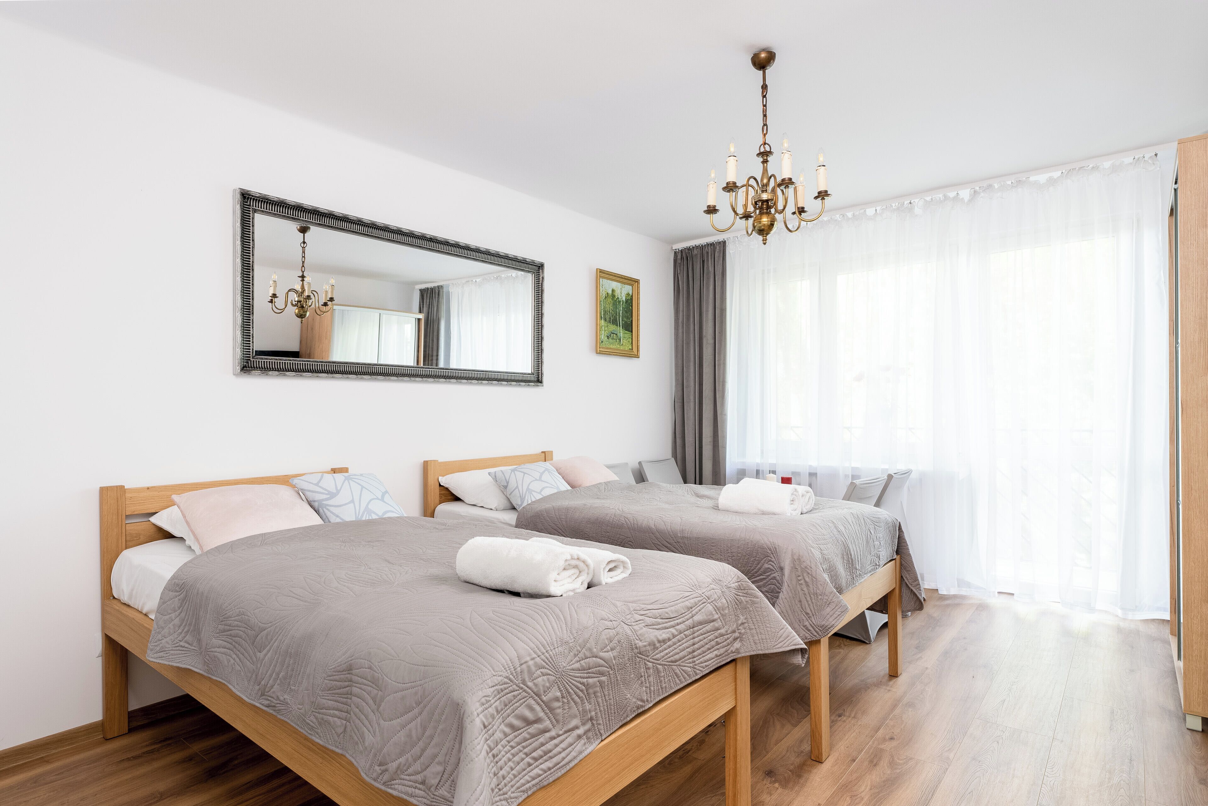 Habitación de hotel con dos camas de madera, un espejo grande, una araña y un amplio ventanal con cortinas de gasa.
