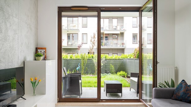 A modern living room with a sofa, a coffee table, a TV, and a sliding glass door leading to a balcony with outdoor furniture and greenery.