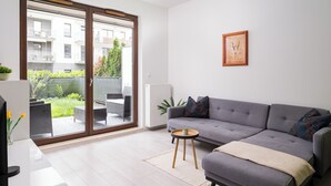 A modern living room with a grey sofa, a small round table, and a sliding glass door leading to an outdoor area with a view of buildings and greenery.