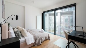 A modern bedroom with a large window, a bed with a beige blanket, a desk with a chair, and a small plant on the bedside table.