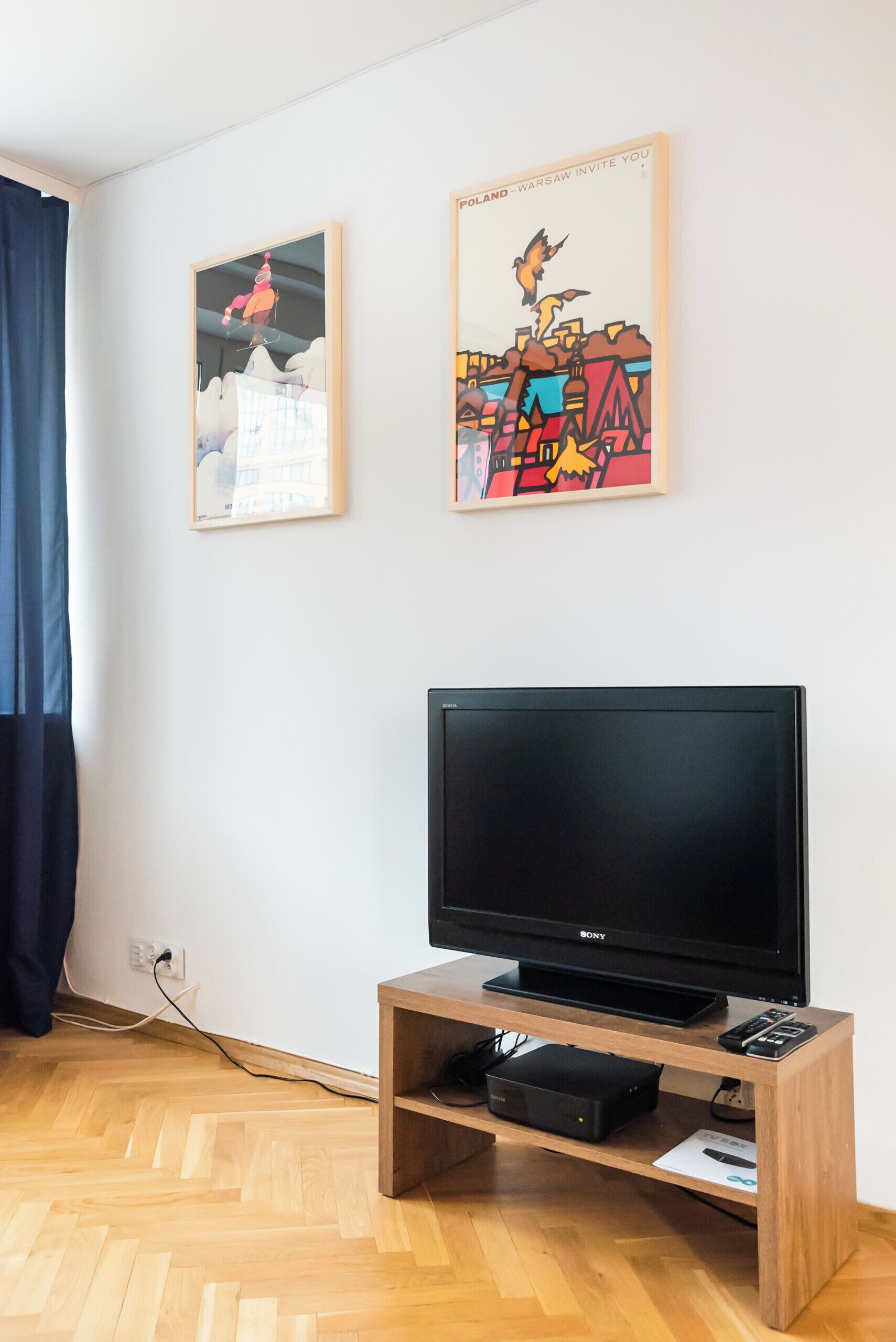 Ein Zimmer mit Holzboden, einem schwarzen Sony-Fernseher auf einem Standfuß, einem kleinen schwarzen Lautsprecher und zwei gerahmten Kunstwerken an der Wand.