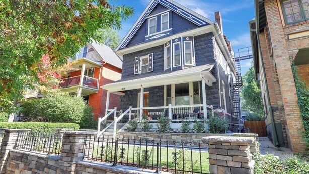 A two-story house with a front porch, a brick chimney, and a stone wall with a metal fence. - Cozy and Chic 1-Bedroom Apartment in Historic Central Denver - Pet Friendly (Denver)