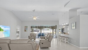 A spacious living room with a large flat-screen TV, a ceiling fan, and a dining area with a glass door leading outside. - Heated Pool, Gulf Access & Dock, Pet Friendly - Villa Canal Breeze - Roelens (Cape Coral)