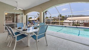A poolside area with a table and chairs, a ceiling fan, and a view of a swimming pool and buildings in the background. - Heated Pool, Gulf Access & Dock, Pet Friendly - Villa Canal Breeze - Roelens (Cape Coral)