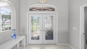 A hallway with a white door featuring frosted glass panels, a white bench with a blue vase, and a chandelier. - Heated Pool, Gulf Access & Dock, Pet Friendly - Villa Canal Breeze - Roelens (Cape Coral)