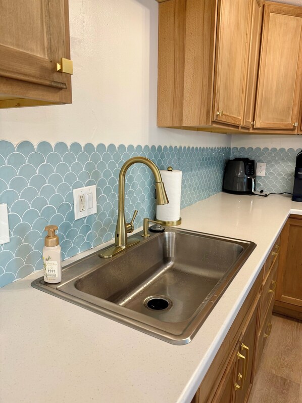 Una cocina con un lavamanos de acero inoxidable, un grifo dorado, armarios de madera, un salpicadero de azulejos azules y una cafetera.