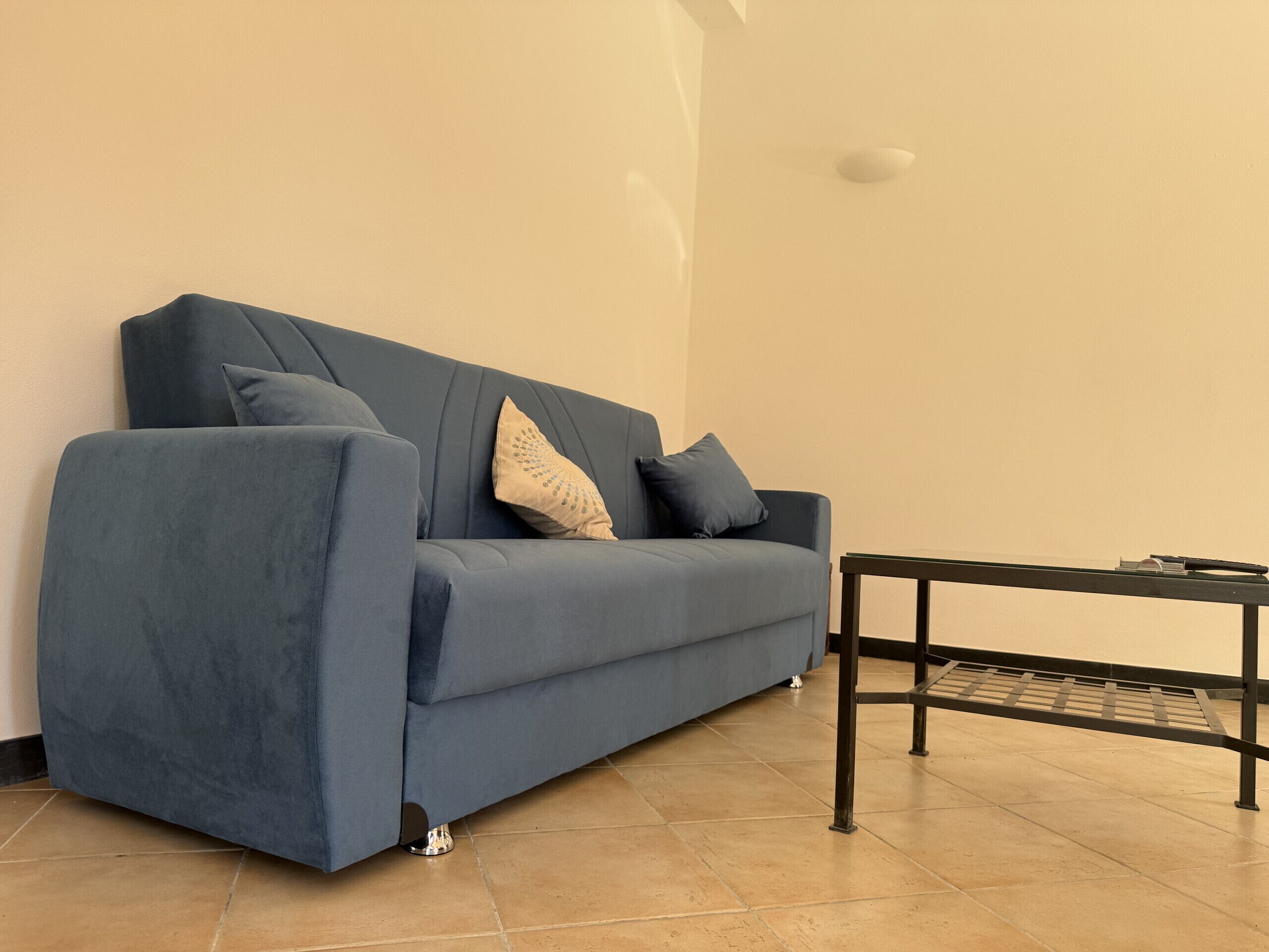 A blue sofa with two pillows against a plain wall, a small black table, and a tiled floor.