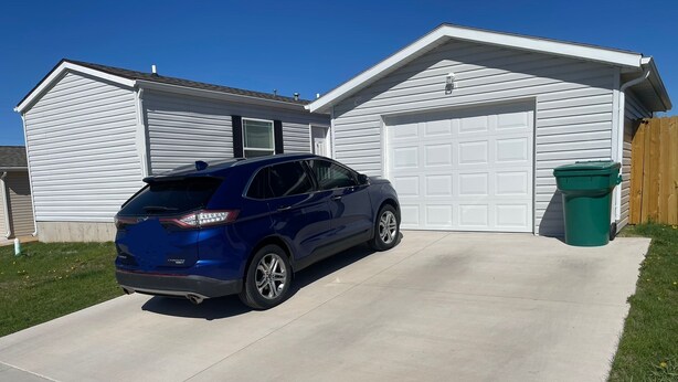 Un VUS bleu est garé devant un garage blanc, avec une poubelle verte à côté.