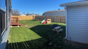 Un jardin avec une terrasse en bois, un toboggan, une petite maison de jeu et une table de pique-nique.